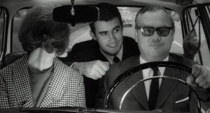 Drei Personen in einem schwarz-weiß Bild aus den 1960ern. Am Steuer Leo Wagner, neben ihm eine Frau und auf der Rückbank ein Mann, der eine Faust macht.